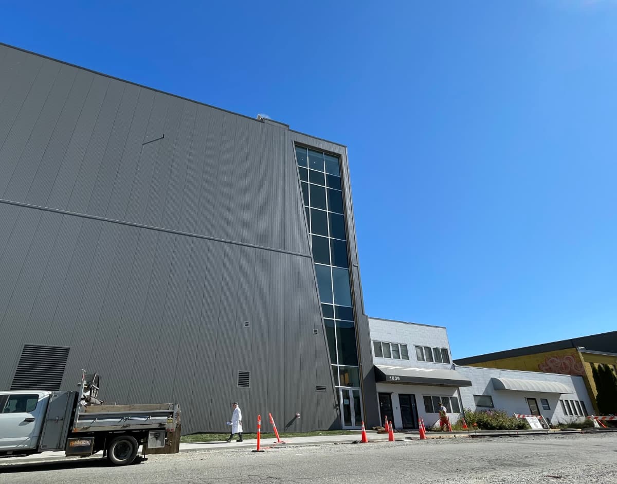 Photograph of part of a big new building, about six storeys tall, and strangely featureless: it looks like a giant metal box, broken only by a handful of vents, with just one column of windows at the corner. It dwarfs the small, old commercial buildings to its right.